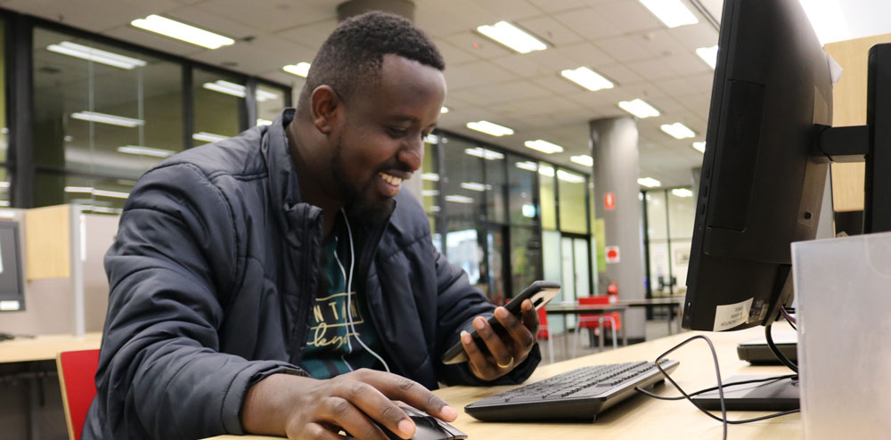 Man smiling and consulting phone while sitting in-front of computer.